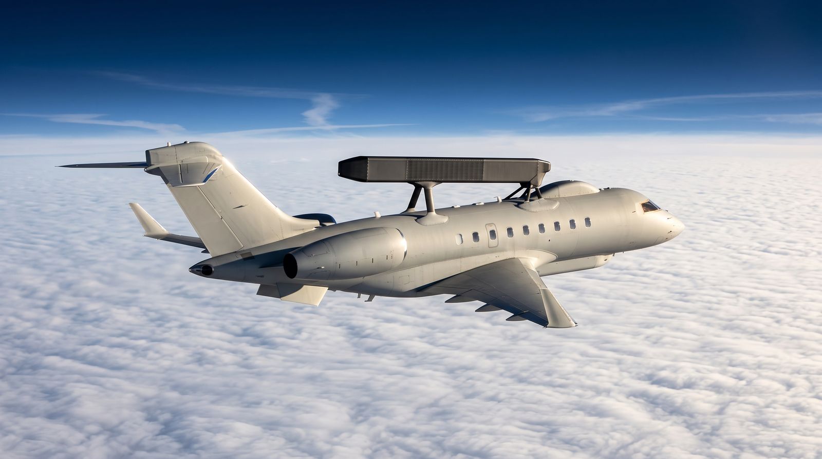 A twin-engine surveillance jet with a dorsal radar spine cruising high above a layer of clouds