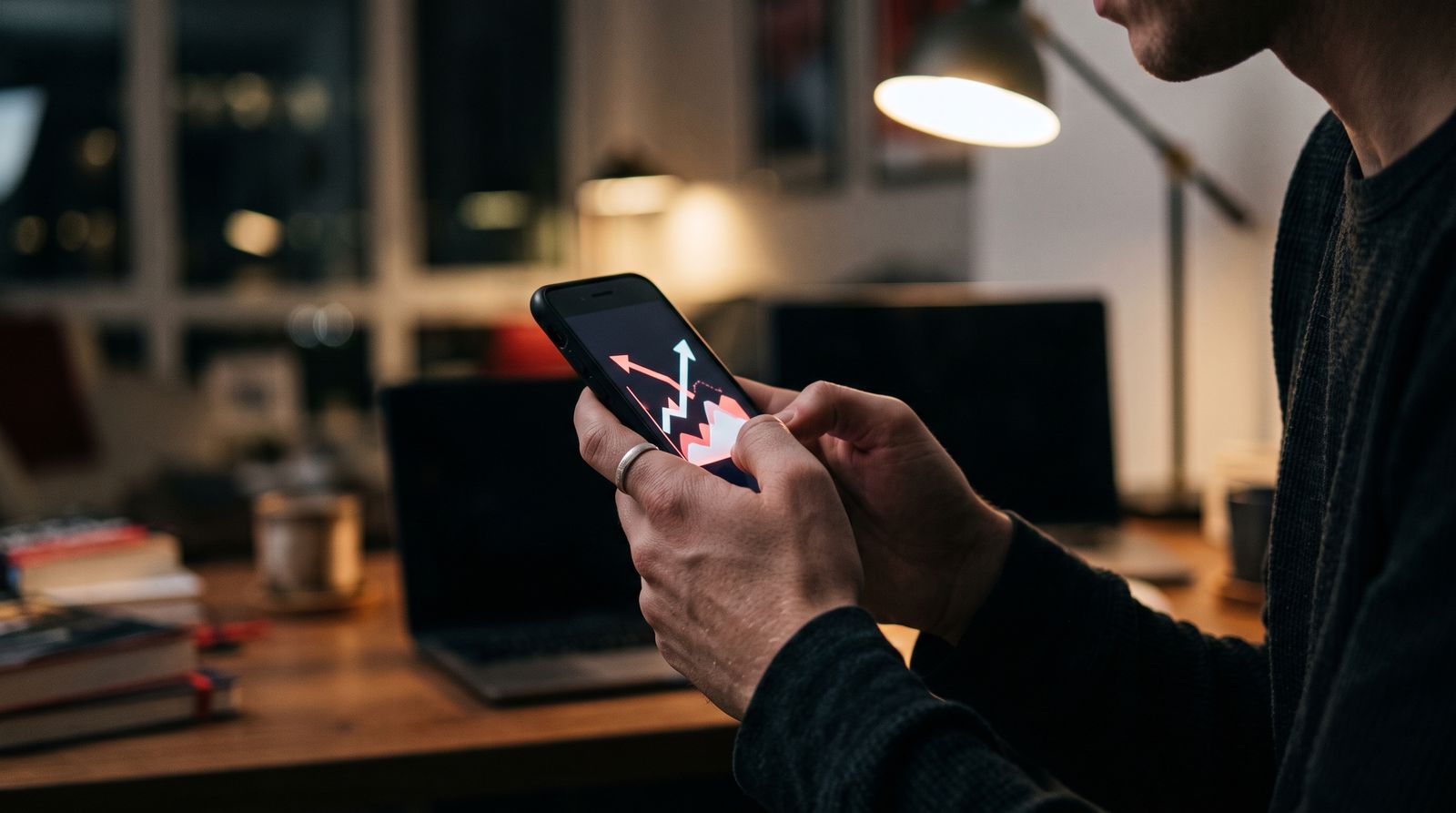 Hands holding a smartphone at a desk at night with abstract market graphics glowing on the screen